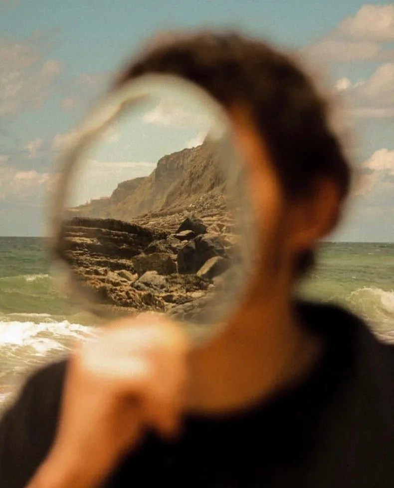 Someone on a beach holds a small mirror up to their face. In it, sharp where everything else is blurred, a sunlit cliff and rocky shore.